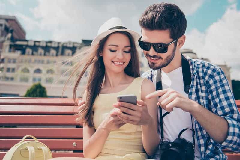 man and woman sitting on park bench and looking at smartphone