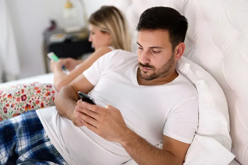 man typing on phone while woman is on the other side of bed