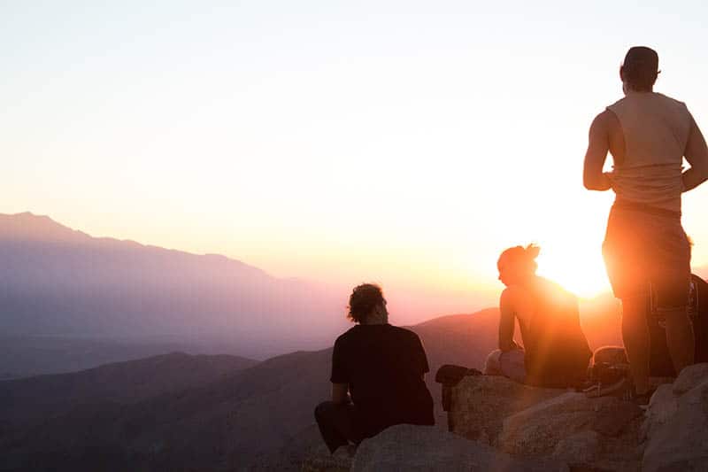 Dois homens e uma mulher sentados nas rochas a ver o pôr do sol