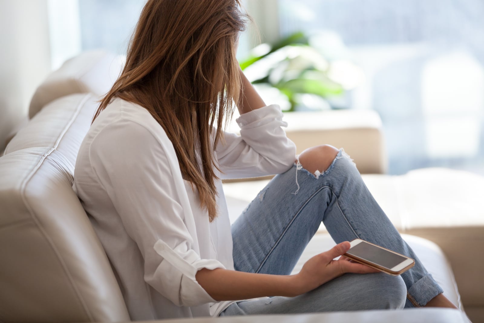 a sad woman with a smartphone in her hand is sitting on the couch