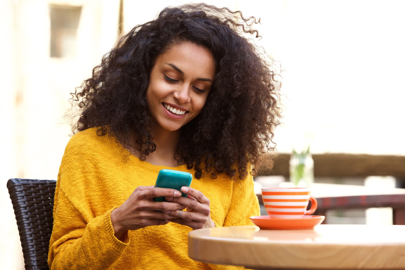 a smiling happy black woman in a yellow sweater sits drinking coffee and texting