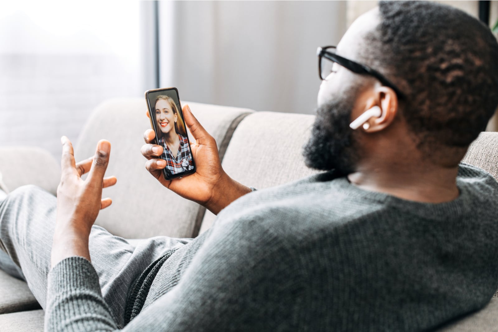 the black man sits on the couch and talks to the smiling blonde over a video call