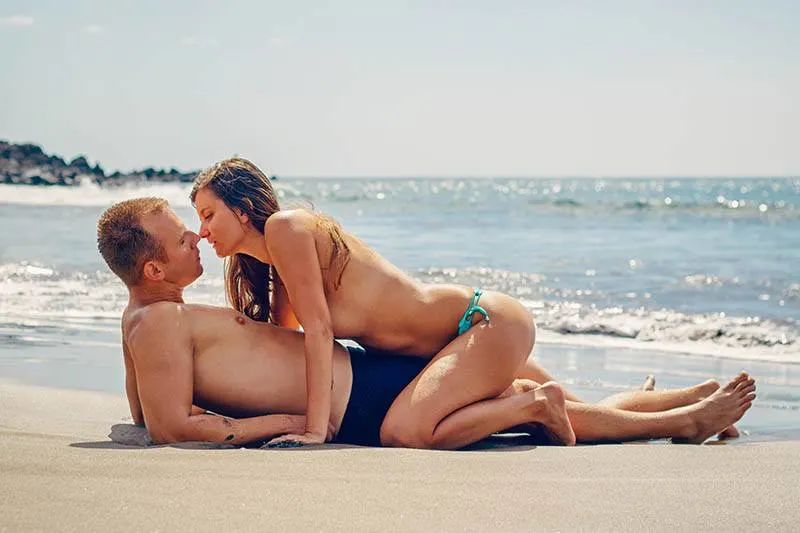 woman in biking sits on man and kisses him while he lying on the beach