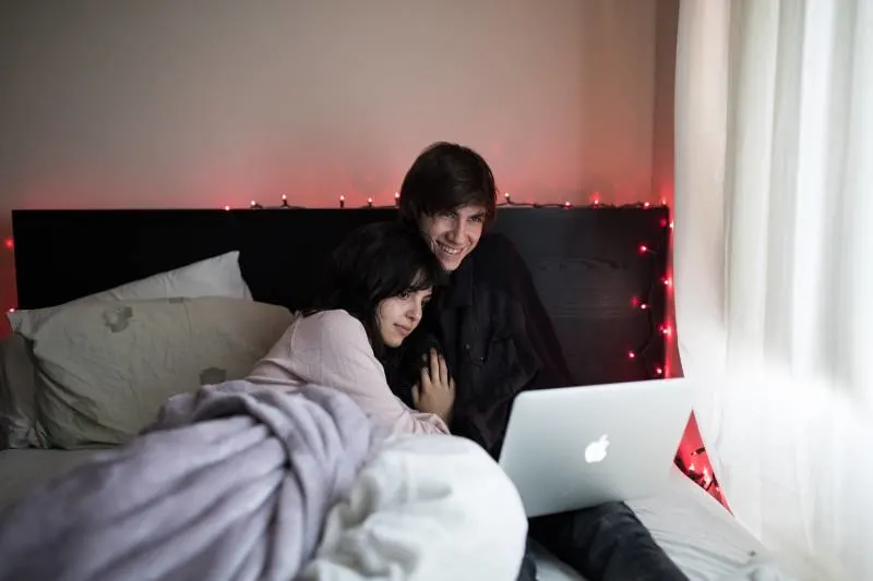 Couple on bed with MacBook Air next to window with white curtain