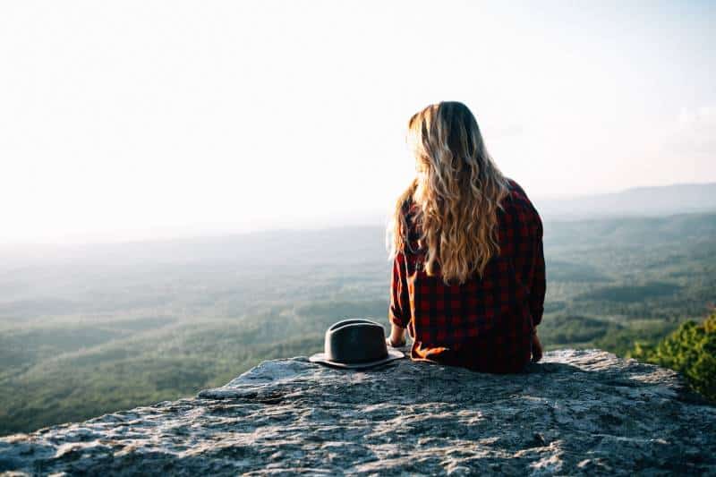 Woman wearing red and black gingham shirt sitting on cliff with hat at his side