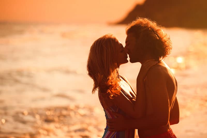 Young woman in bikini kissing her boyfriend on beach at sunset. Lovely couple