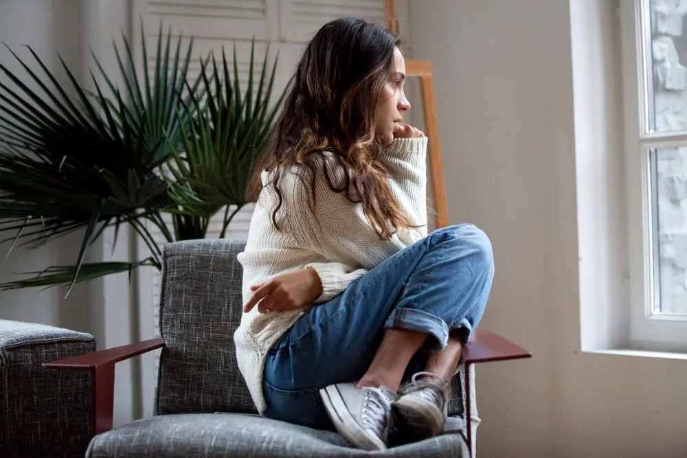 a sad girl in a white sweater, pants and sneakers sits in an armchair