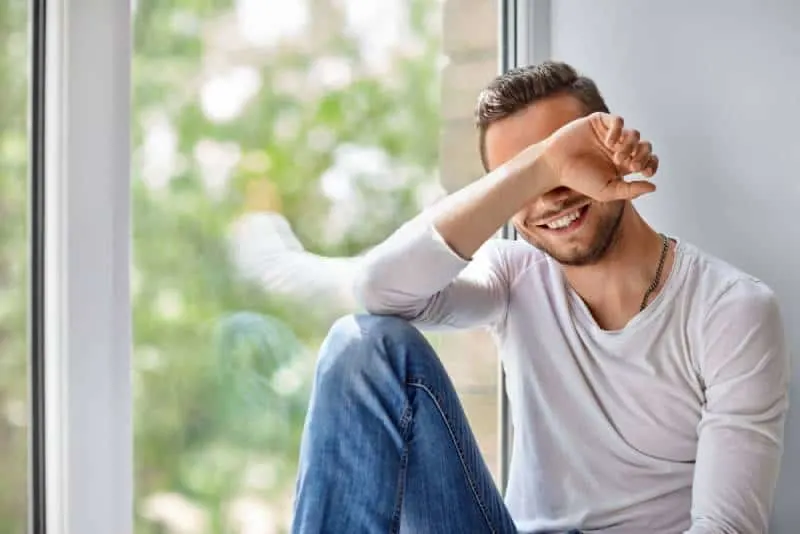 smiling shy man close face with hand while sitting beside window