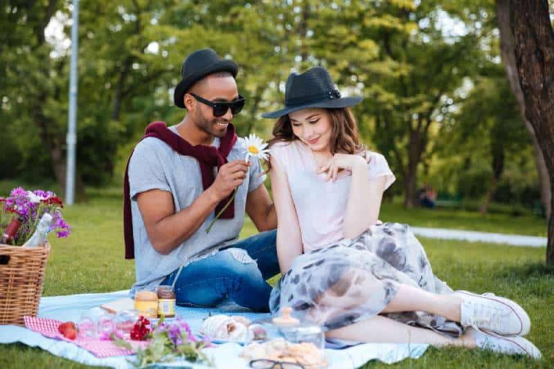 joven sonriente dando flor a su tímida novia en picnic