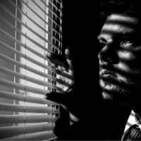 a man from a dark room peeks through the blinds