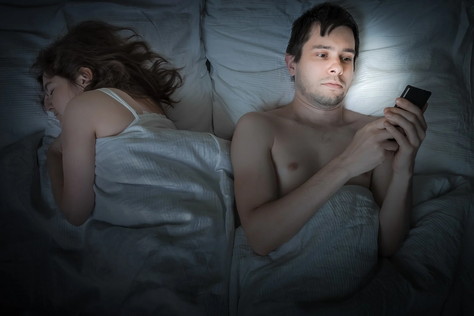Young man is lying with girlfriend in bed and texting with phone at night