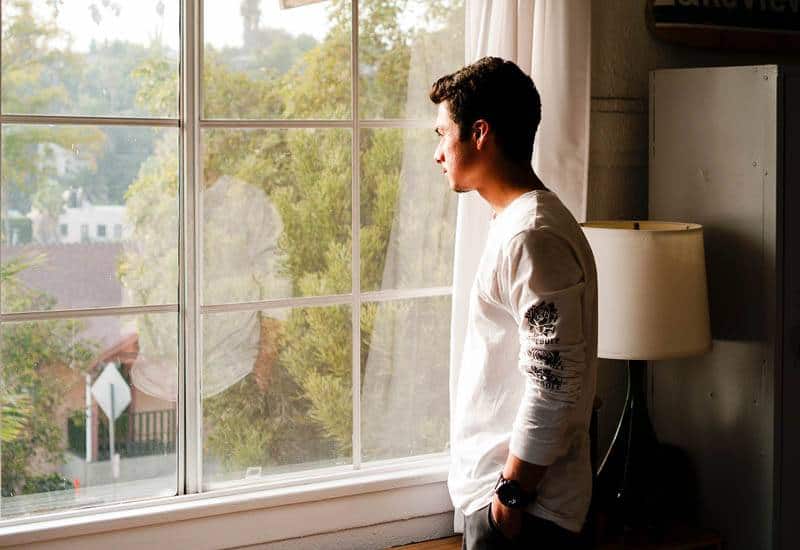man in white tshirt stands beside window and looking out