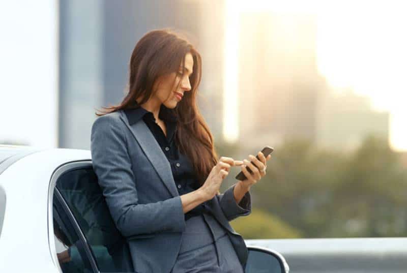 donna d'affari sorridente che scrive sul suo telefono mentre si appoggia all'auto all'aperto