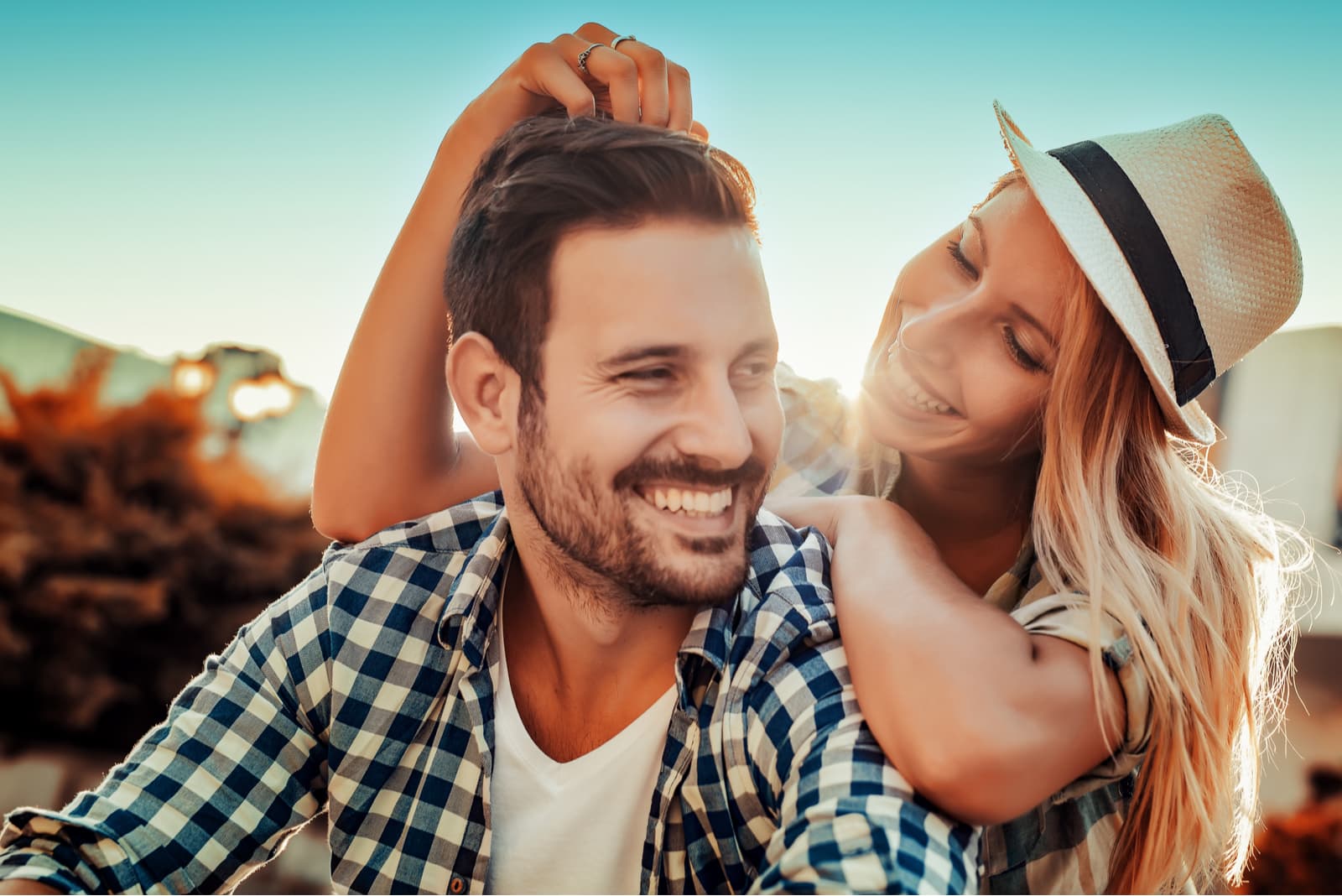 uma mulher de cabelo loiro e chapéu toca na cabeça de um homem e ri-se