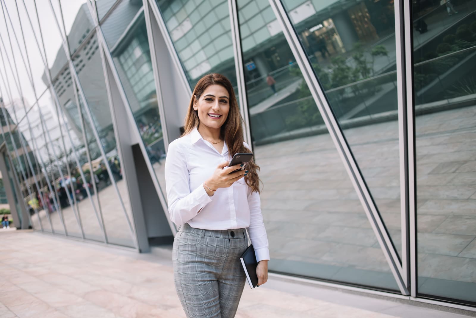 a business smiling woman in a white shirt standing outside with a cell phone in her hand