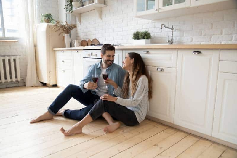 coppia romantica seduta sul pavimento della cucina che beve vino e si guarda sorridendo