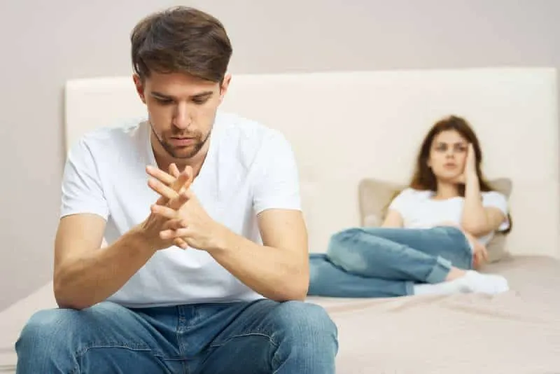 upset man sitting on bed while his girlfriend lying on bed
