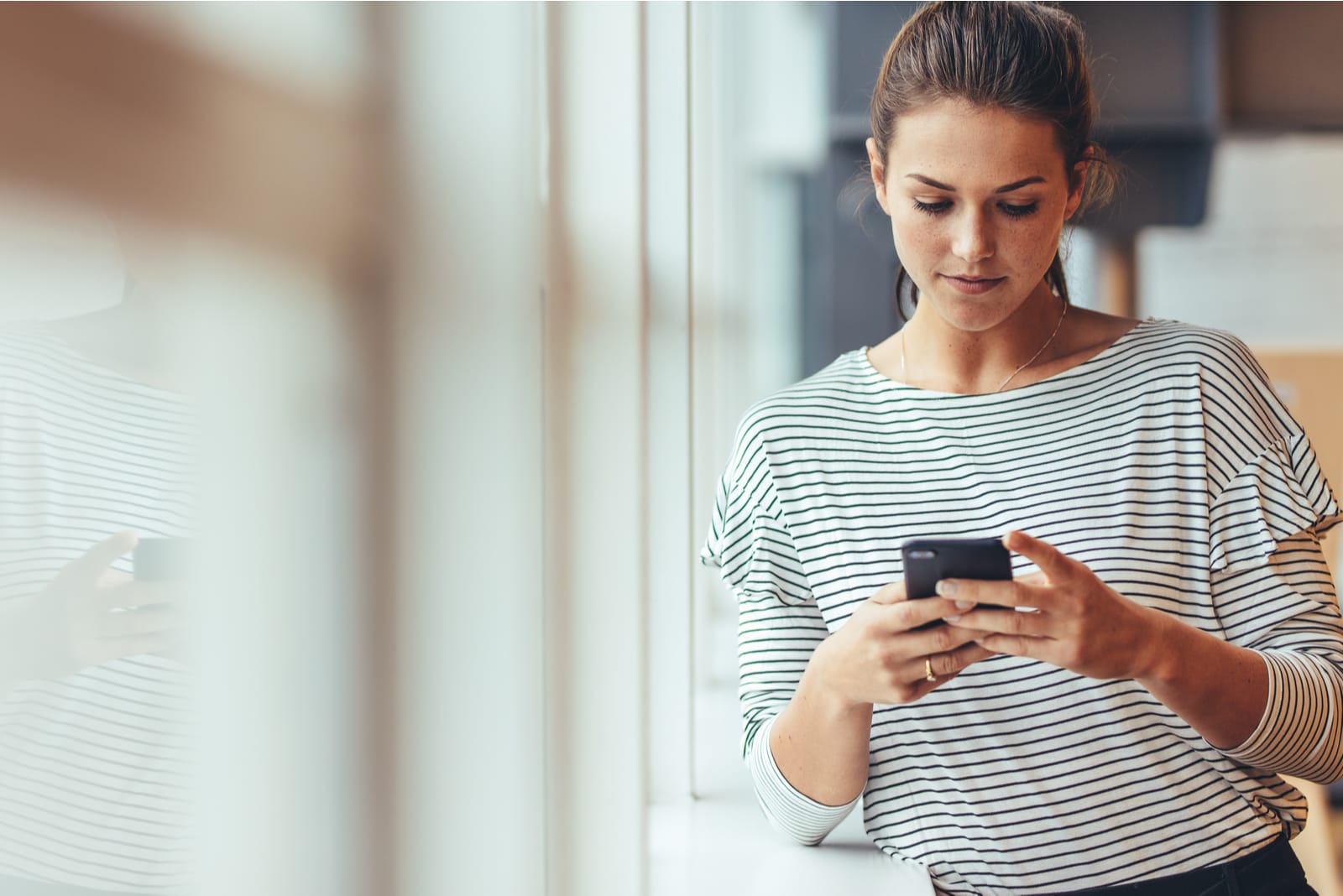 woman standing beside a window holding mobile phone