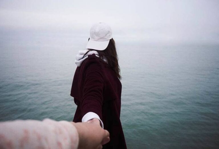 man holds woman hands while she facing sea