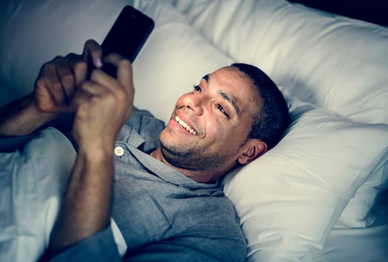 man in pajamas lying on bed and typing on his phone