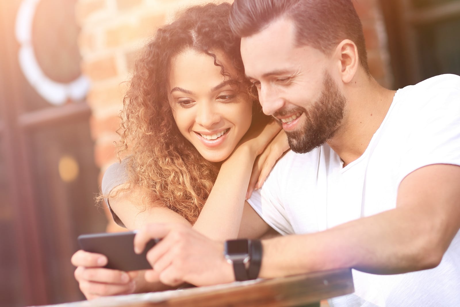 um casal feliz e apaixonado sentado no terraço de um café e a utilizar um smartphone