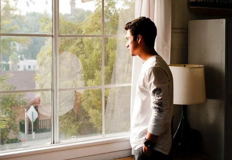 man in white t-shirt stands beside window and looking out