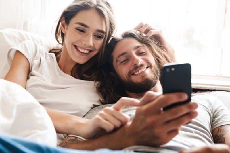 pareja sonriente en casa miente usando el móvil