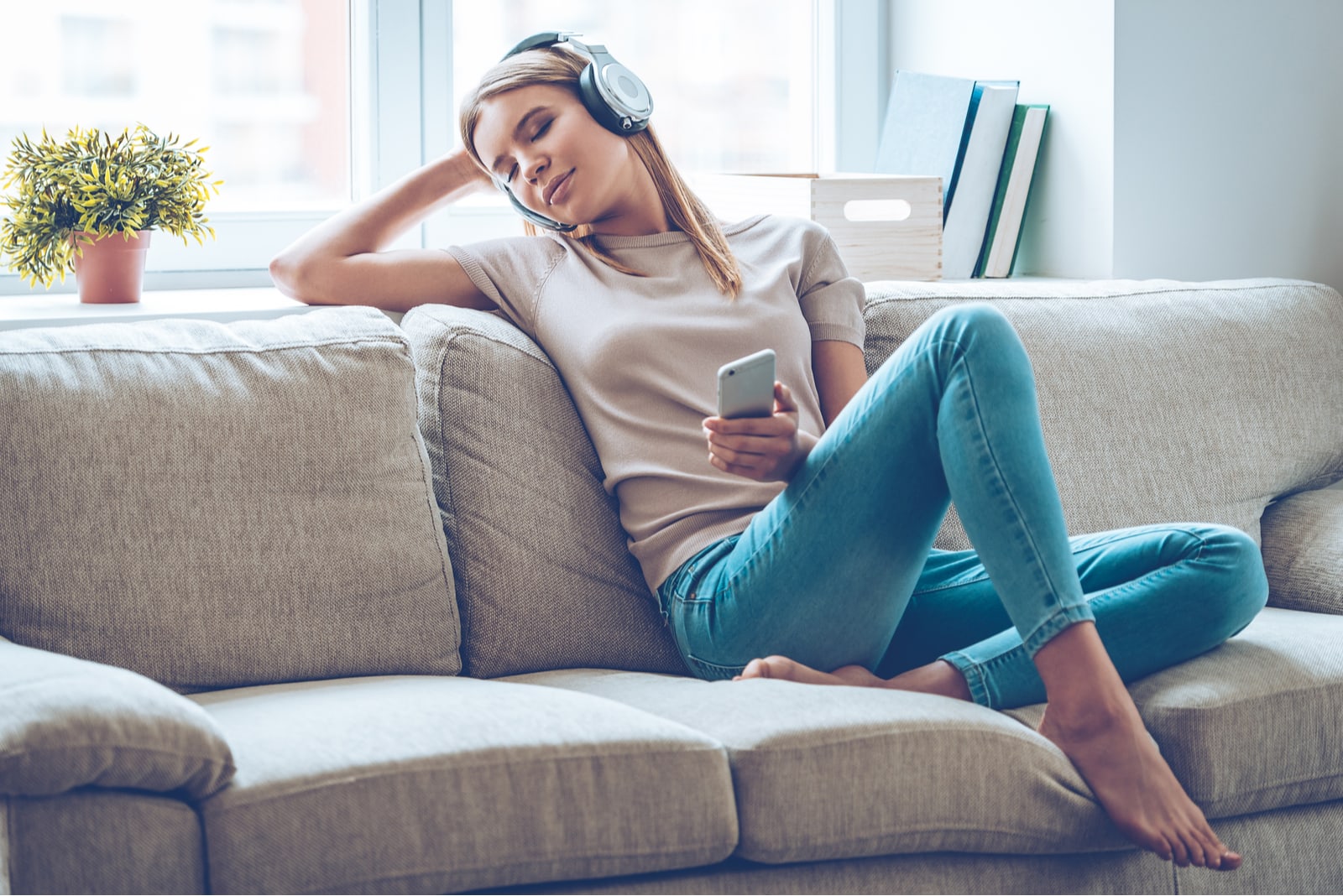 a beautiful blonde sits on the couch and listens to music through headphones on her smartphone