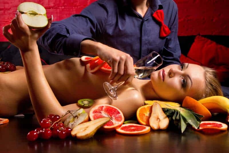 mulher nua decorada com frutos enquanto um homem segura um copo de champanhe