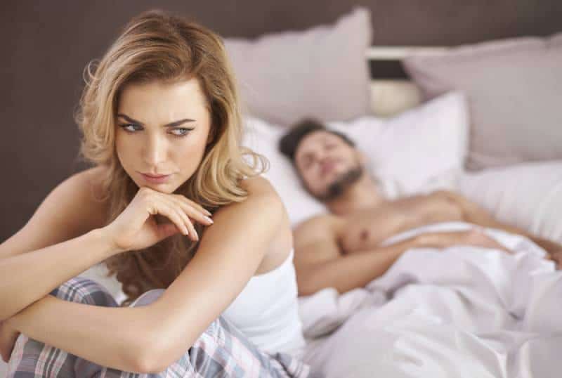 mujer pensativa sentada en la cama mientras el hombre acostado