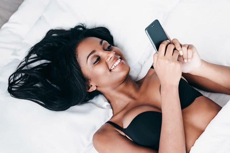 giovane donna sorridente sdraiata sul letto che scrive sullo smartphone