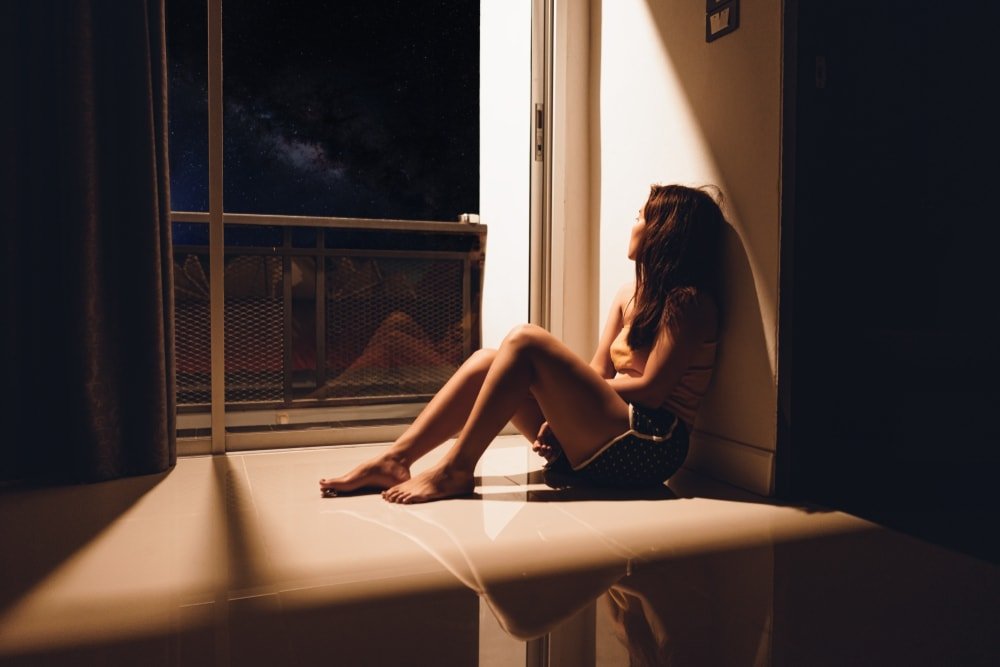 a depressed brunette sits on the floor by the window in the dark