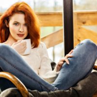 an attractive red-haired girl sits in an armchair and drinks tea