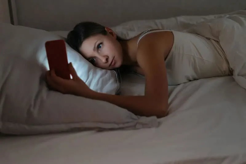 woman lying on white pillow and looking at her phone