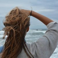 back view of woman touches her hair while standing in front of sea