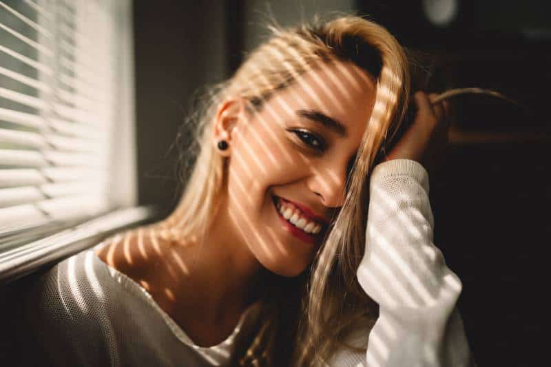 selective focus photography of the smiling woman holding her hair next to blinds