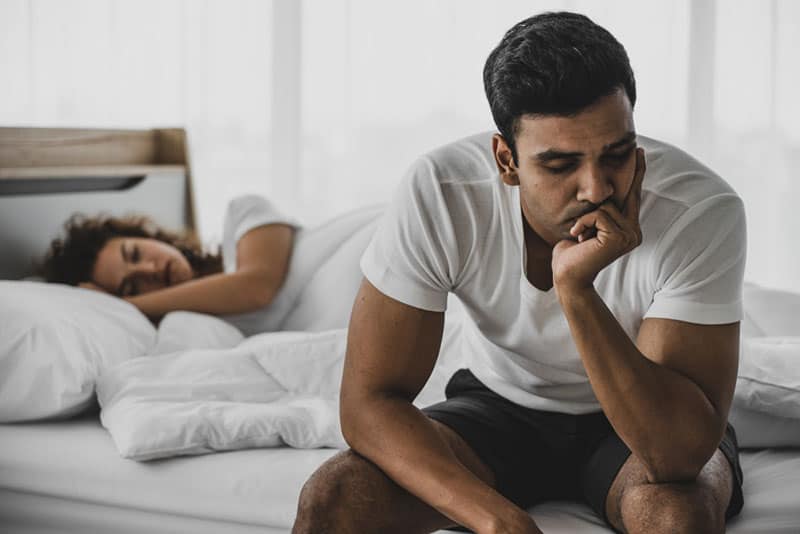 worried man sitting on bed while woman sleeping