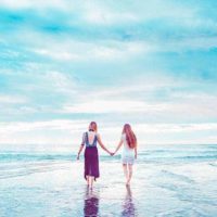 back view of two female friends holding hands while walking on the beach
