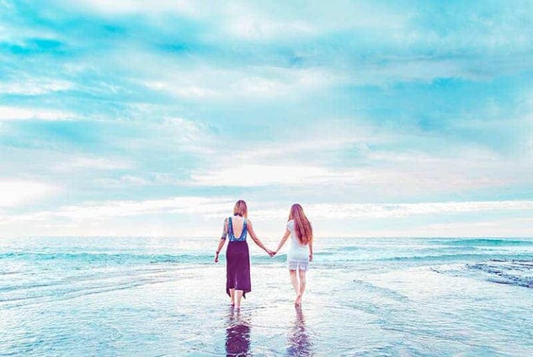 back view of two female friends holding hands while walking on the beach