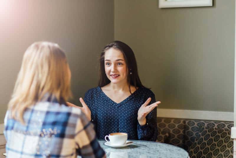 Cheerful woman shares the news with her friend while drinking cappuccino in a cafe