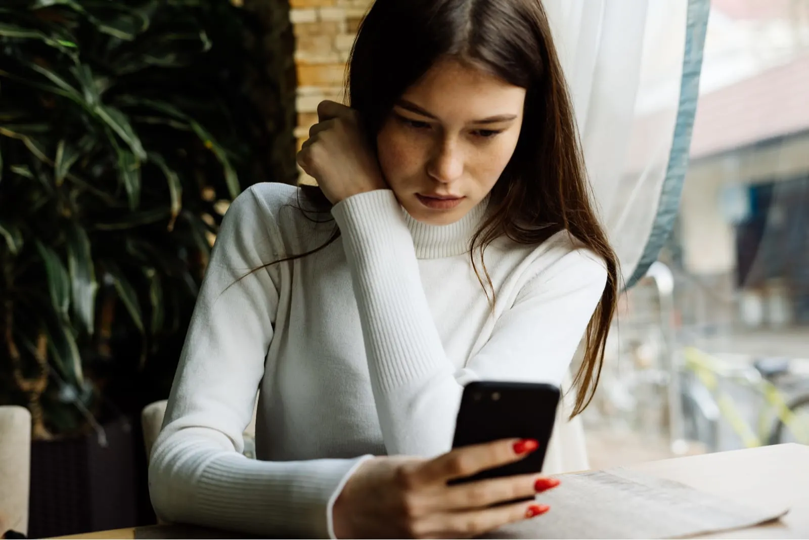 a serious woman in a white sweater is sitting in a cafe by the window and using a smartphone