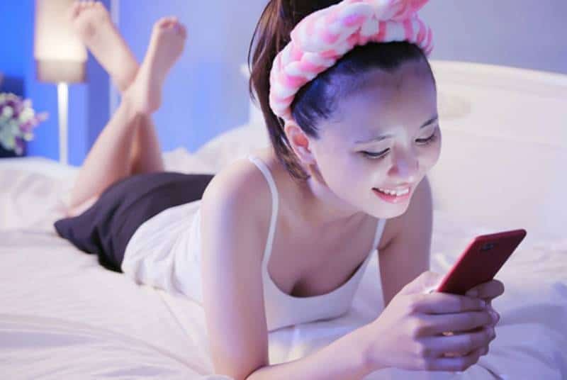 donna sorridente sdraiata sul letto che scrive sul suo telefono