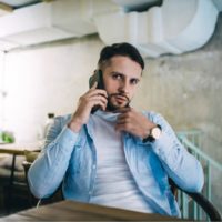 a serious man in a blue shirt is sitting in a cafe and calling someone on his cell phone