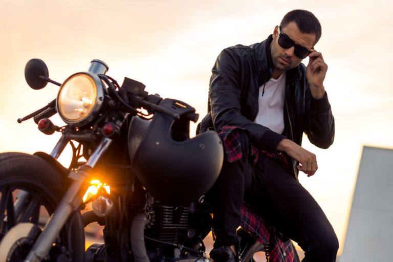 a handsome man in a black leather jacket with reyban sunglasses sits on a motorcycle