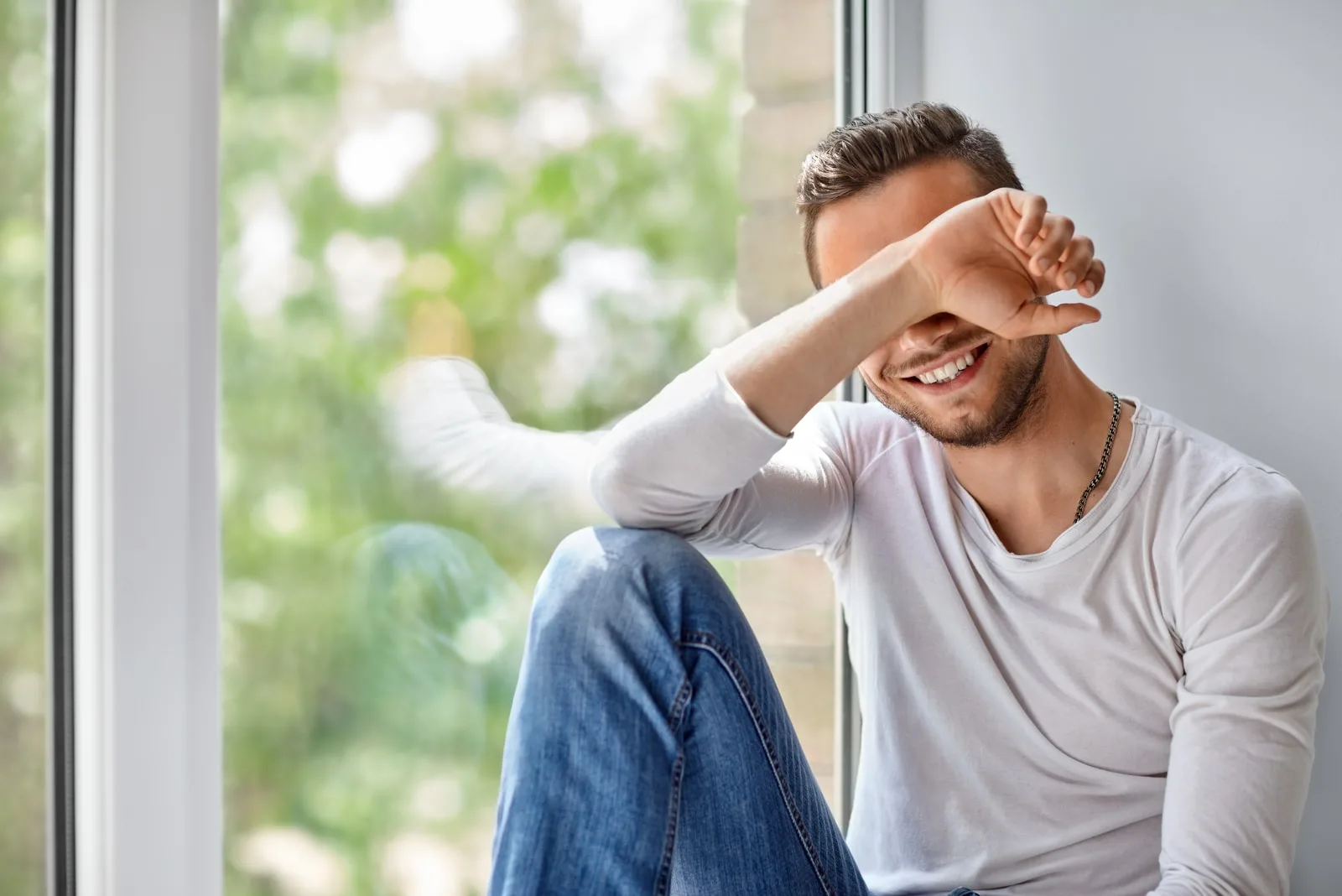 Smiling shy man close face with hand sitting on window sill