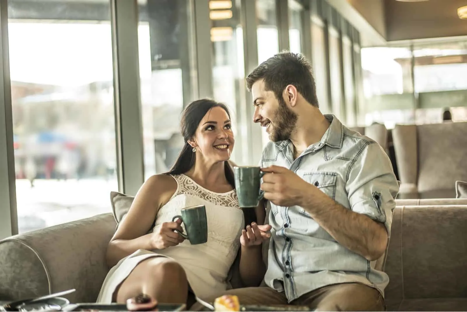 a smiling man and woman sit on the couch drinking coffee and talking