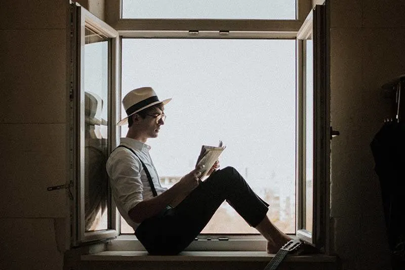 man reading book while sitting on window
