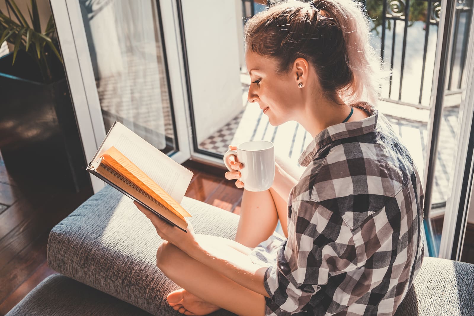 bonita mujer sentada junto a una ventana abierta bebiendo café y leyendo un libro