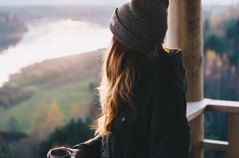 woman wearing hat and black jacket holds cup of tea while looking in nature