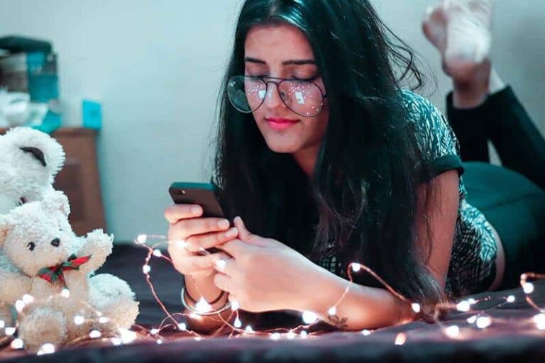 girl wearing eyeglasses lying on bed and typing on her phone